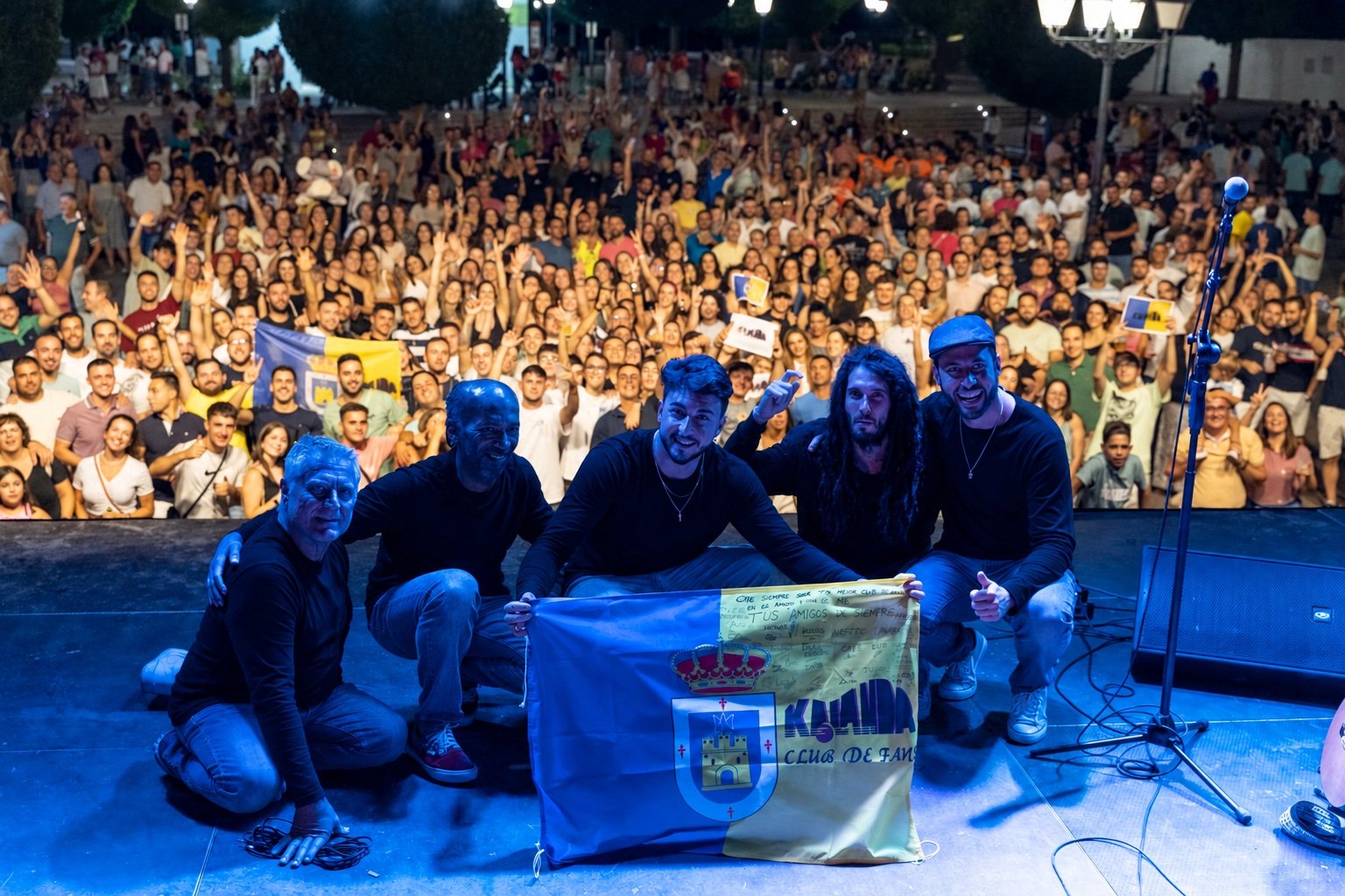 Grupo Kalanda posando en el escenario con el público y la bandera de su club de fans